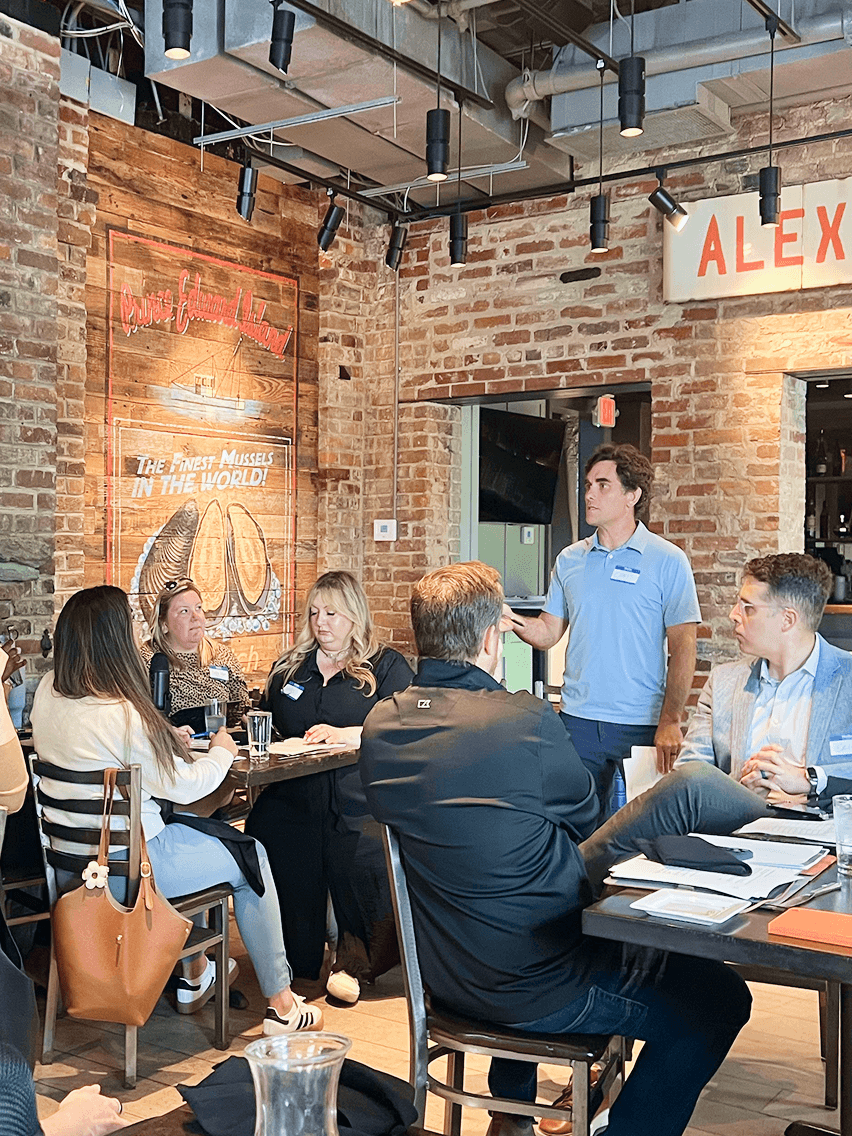 Group of people seated at tables in a brick-walled restaurant, one man standing and speaking to the group.