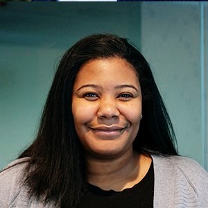Smiling woman with long brown hair wearing a sleeveless black wrap dress against a black background.