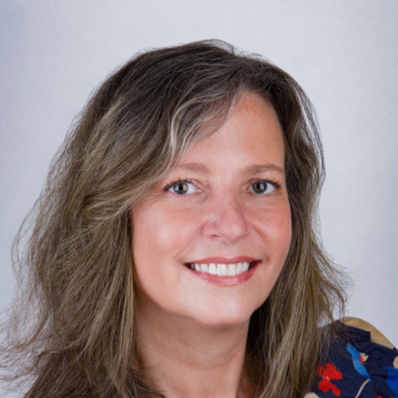 Smiling woman with long brown hair wearing a sleeveless black wrap dress against a black background.