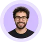 Smiling man with curly hair, glasses, and a beard in front of a purple circular background.