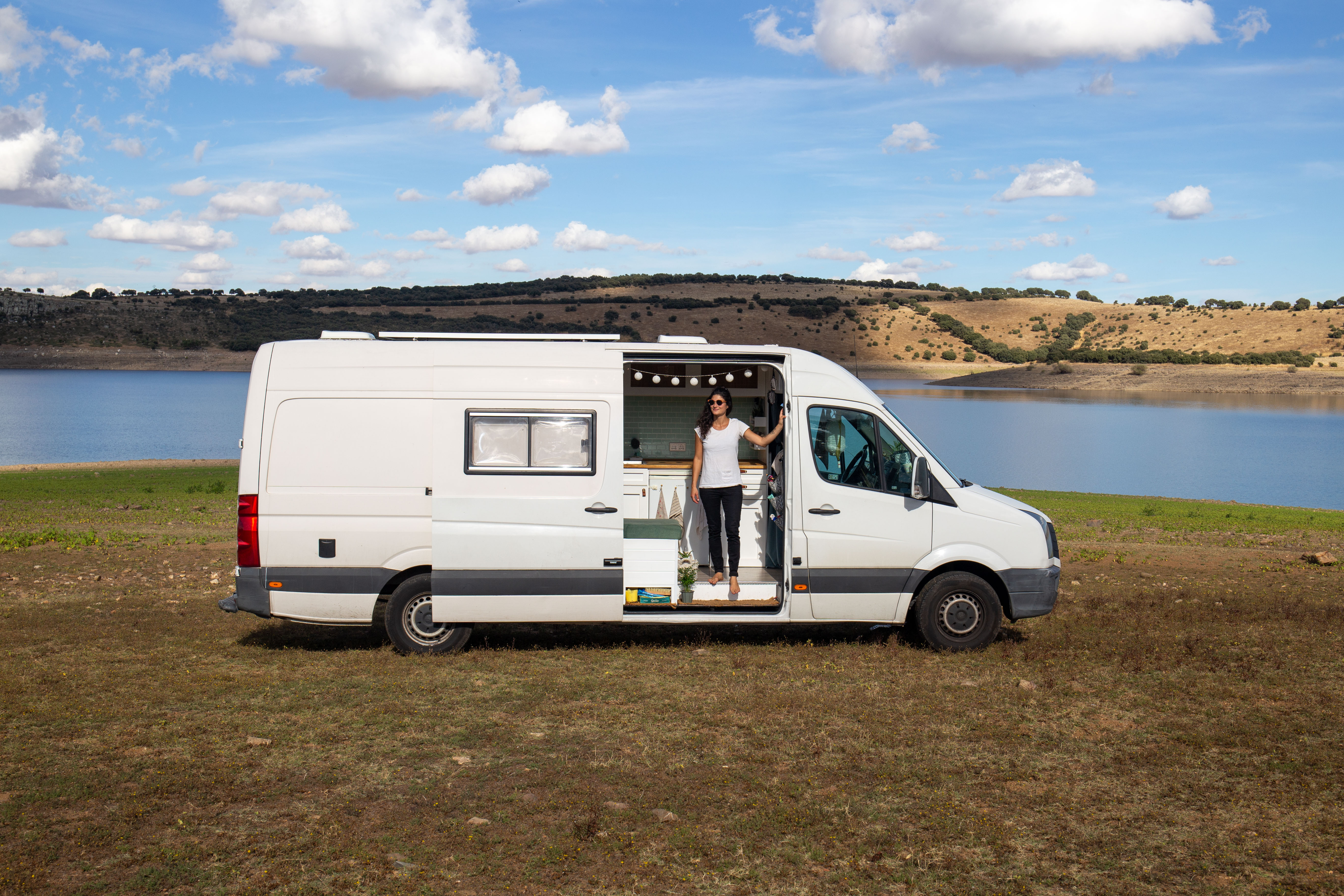 Woman standing barefoot in the open sliding door of a white camper van parked on a grassy area near a lake with hills in the background under a blue sky with clouds.