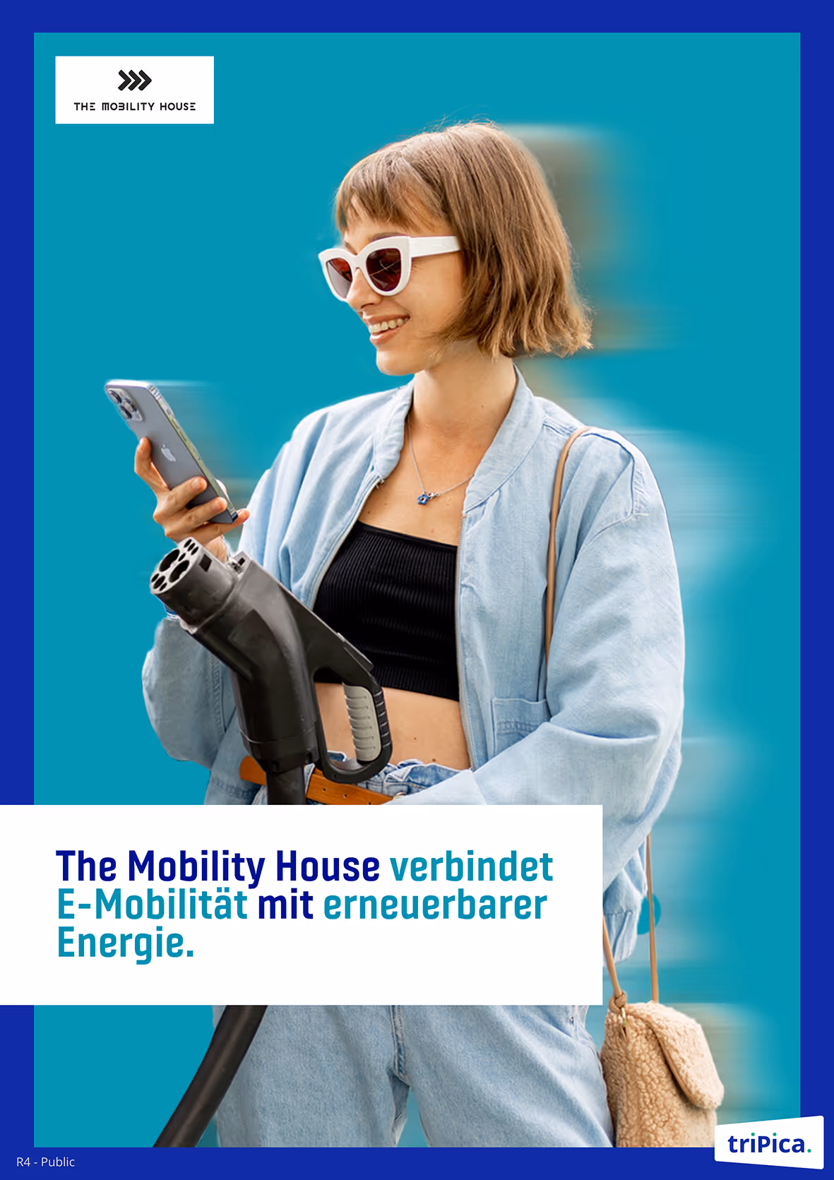 Young woman wearing white sunglasses and a light denim jacket holding an electric vehicle charging plug and looking at her smartphone.