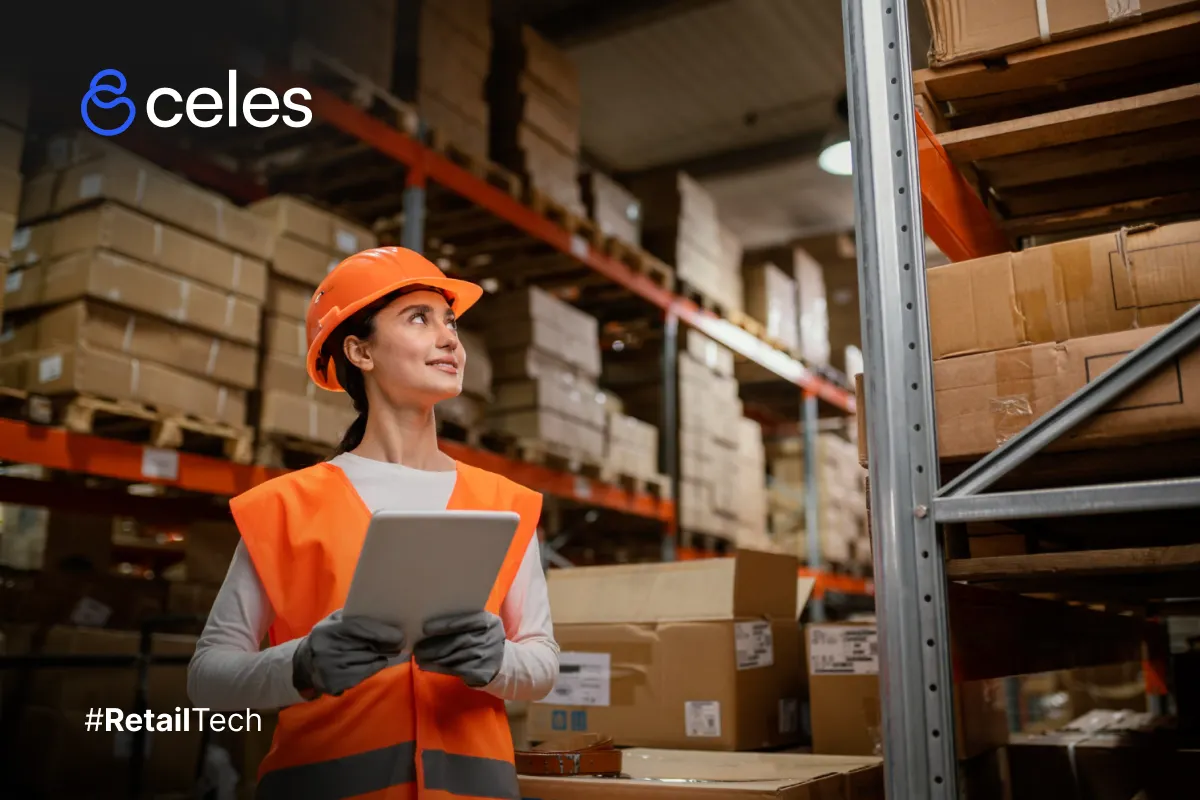 Mujer sonriente con casco naranja y chaleco reflectante, sosteniendo tablet blanca con guantes grises en almacén lleno de estanterías naranjas y cajas de cartón apiladas