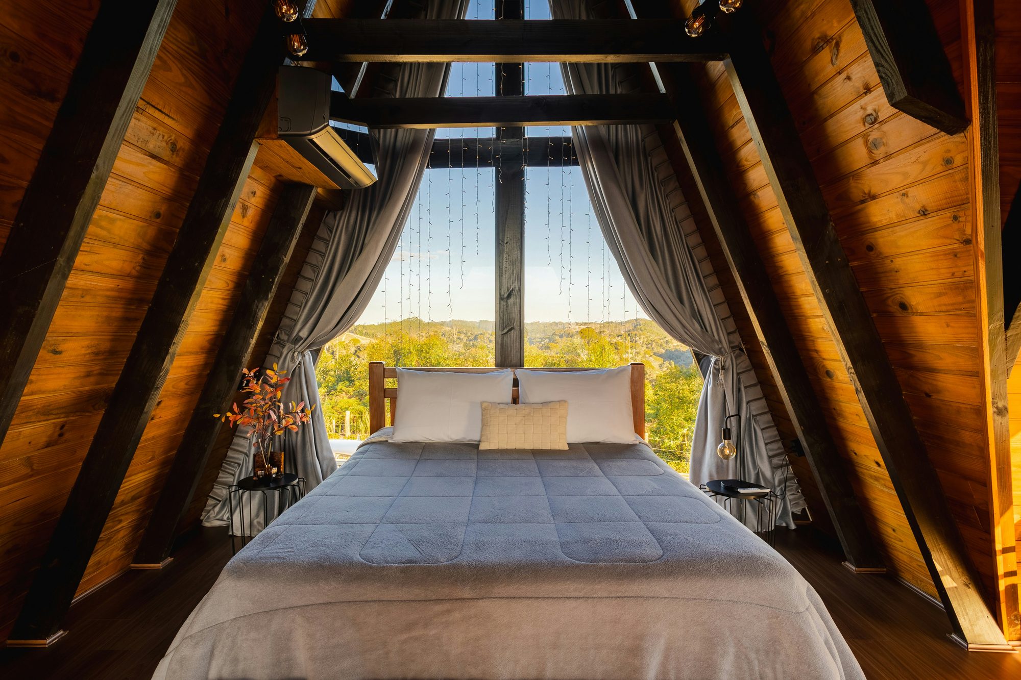 A-frame cabin bedroom with curtains framing a sunset view over wooded hills