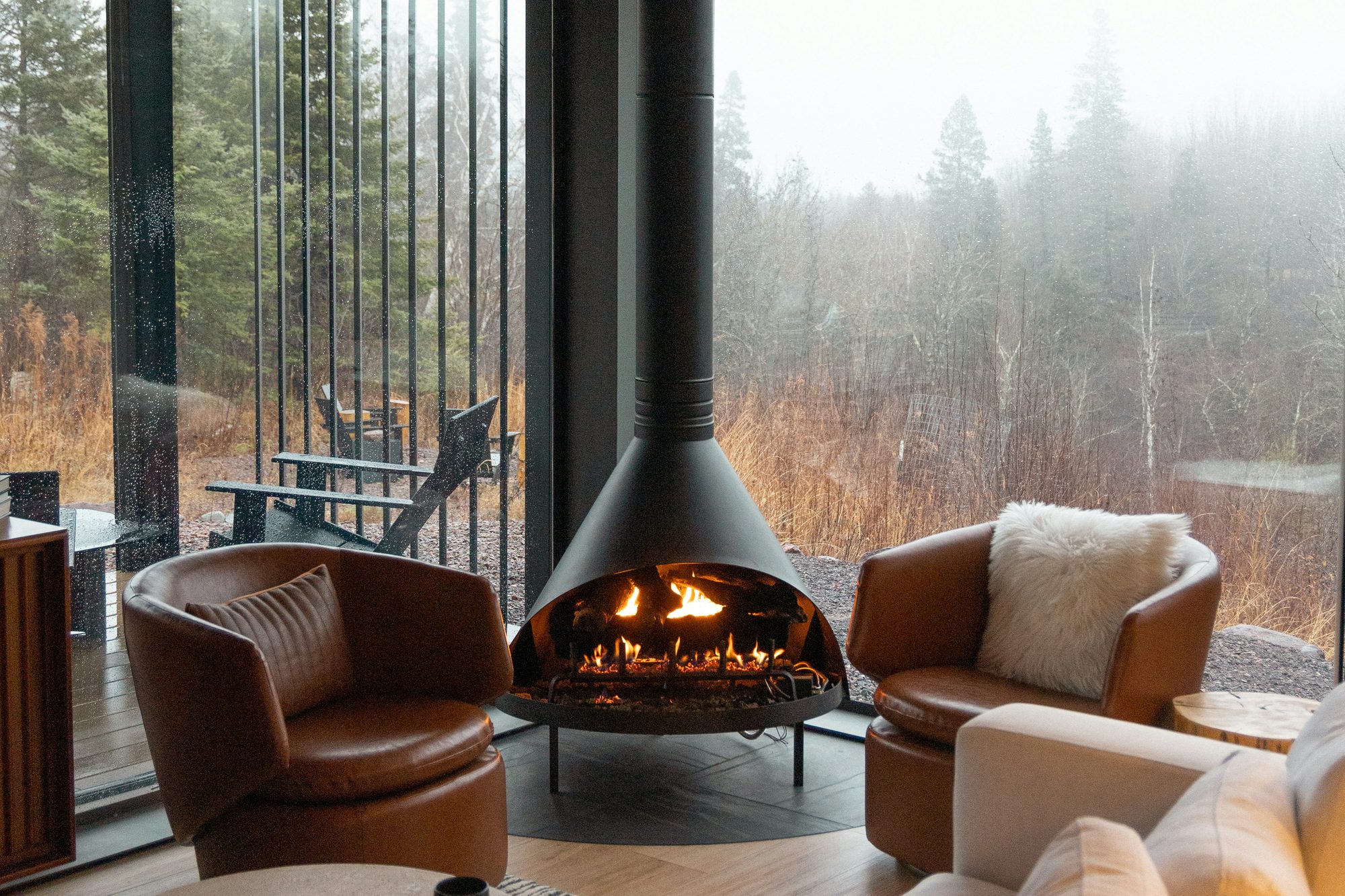 Hanging fireplace between two leather chairs, with floor-to-ceiling windows onto a misty forest