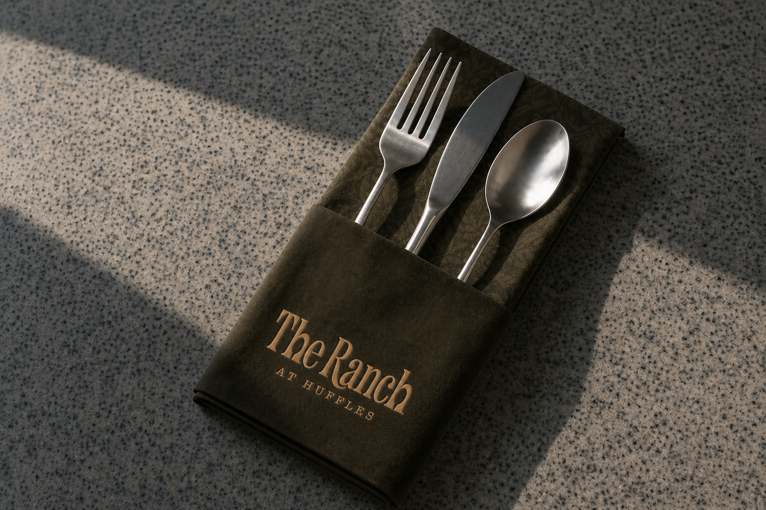 Dark linen napkin embroidered with The Ranch at Huffles logo, holding a fork, knife and spoon