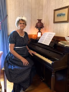 Donna Chaff as Lady Maria Chaffington, in a navy blue patterned dress, sitting at an antique piano with sheet music on the stand