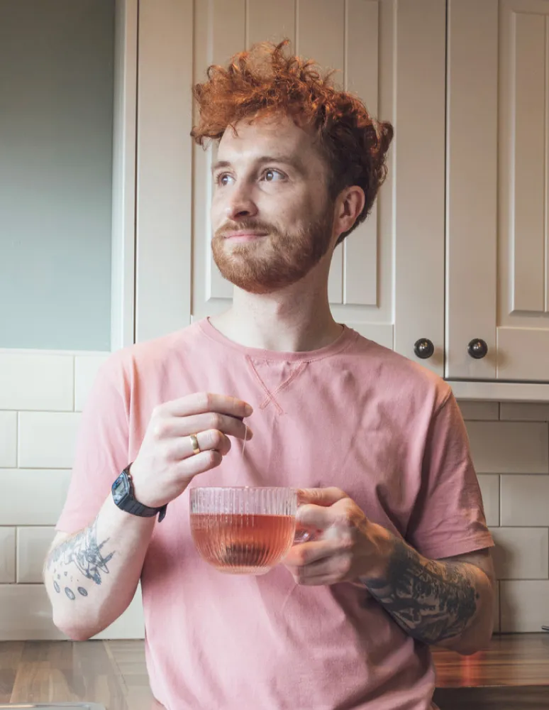 Person with curly red hair holding a glass mug of tea in a kitchen.