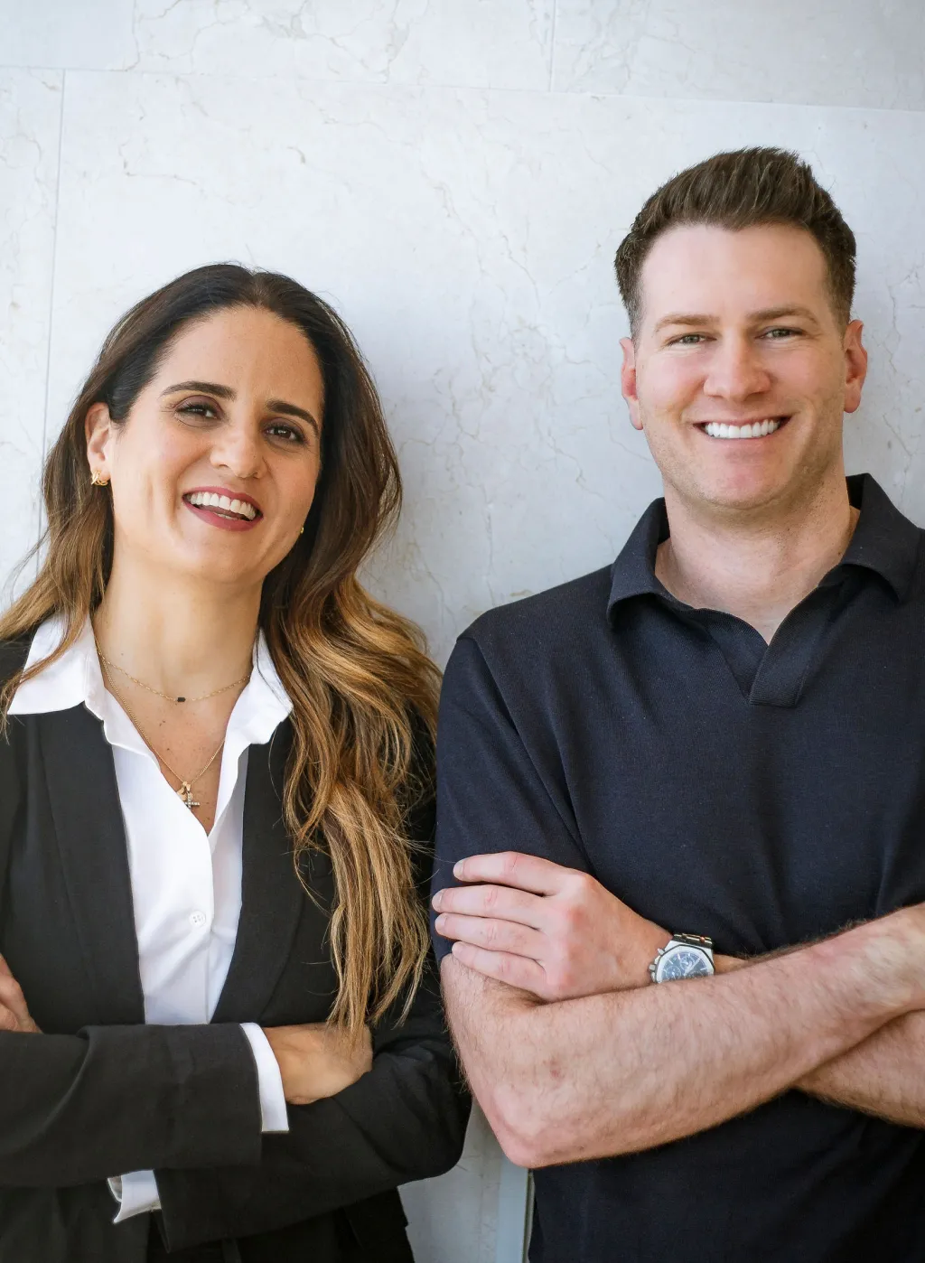 Two professionals smiling with arms crossed, standing against a light stone wall