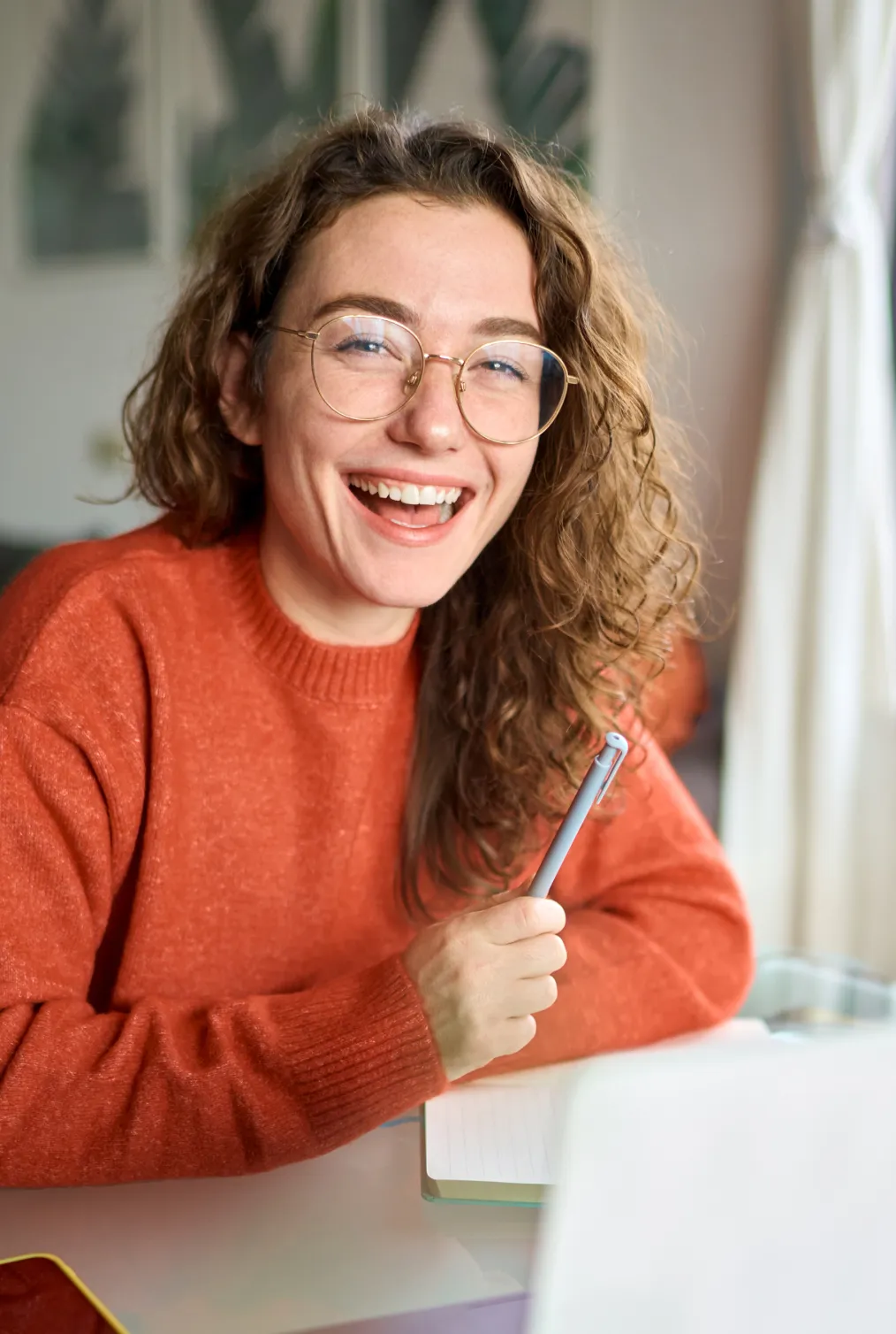 Smiling woman wearing glasses, holding a pen while seated at a desk