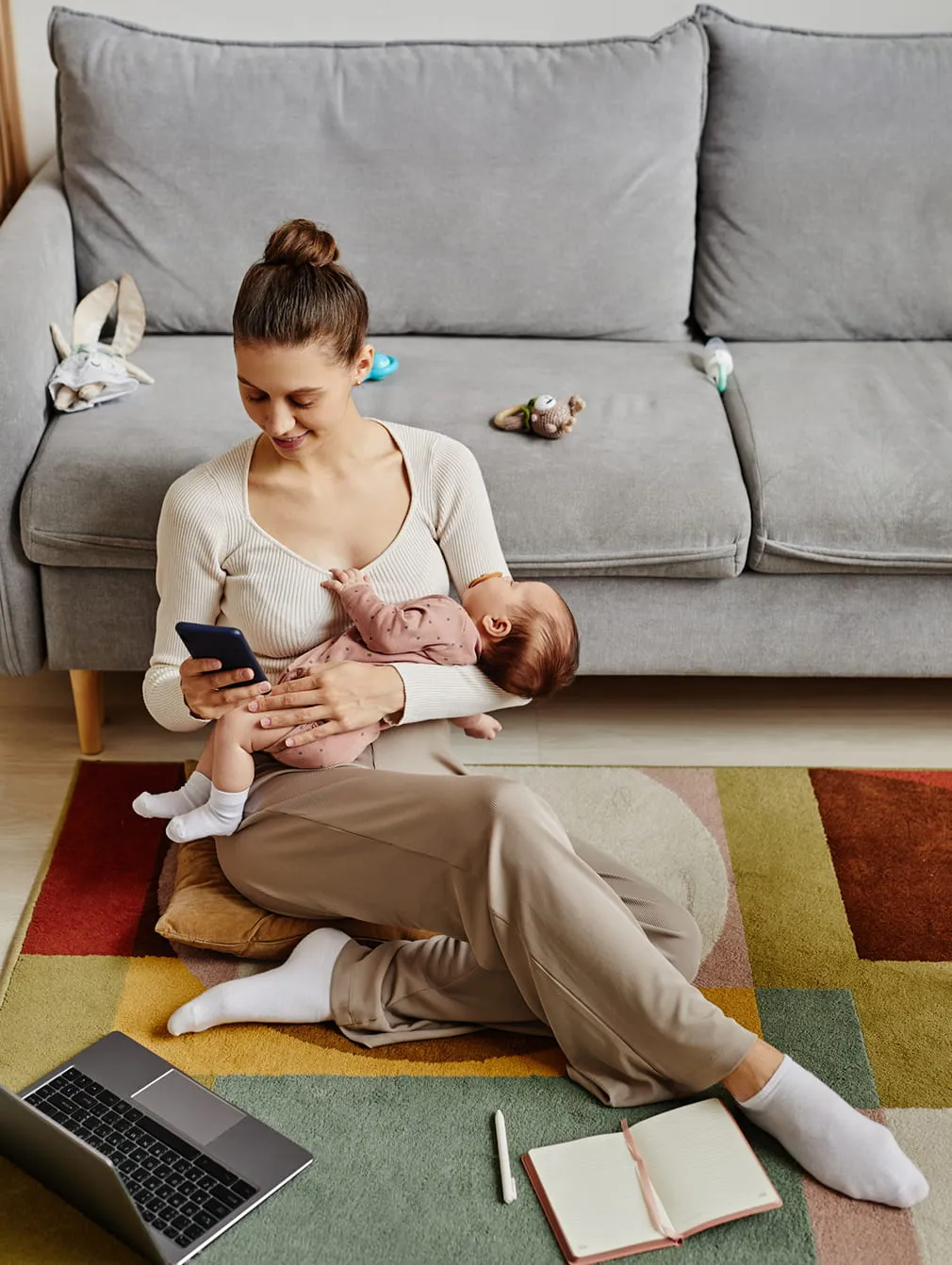 Mother sitting on a floor cushion at home while holding and nursing a baby, looking at her phone, with a laptop and notebook nearby on a colorful rug.