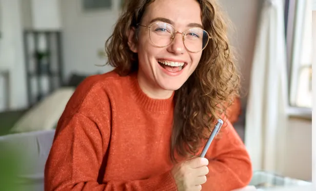 Smiling woman wearing glasses, holding a pen while seated at a desk