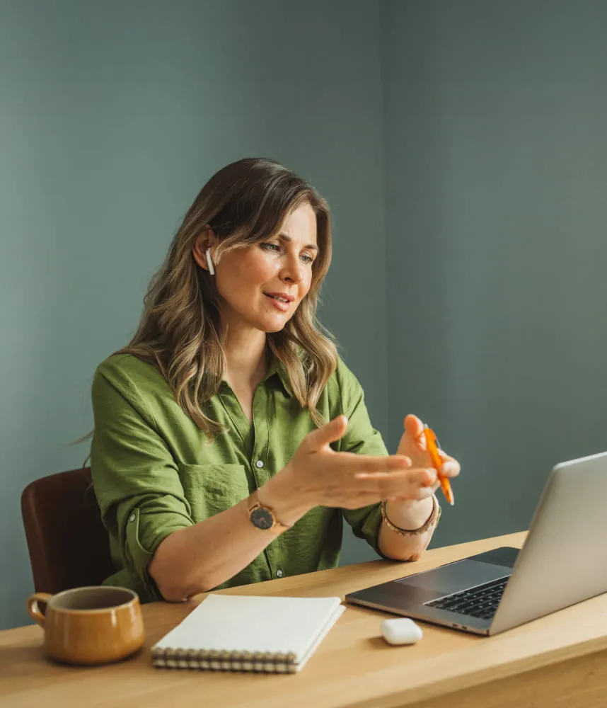 Woman wearing wireless earbuds talking on a video call at a desk with a laptop and notebook