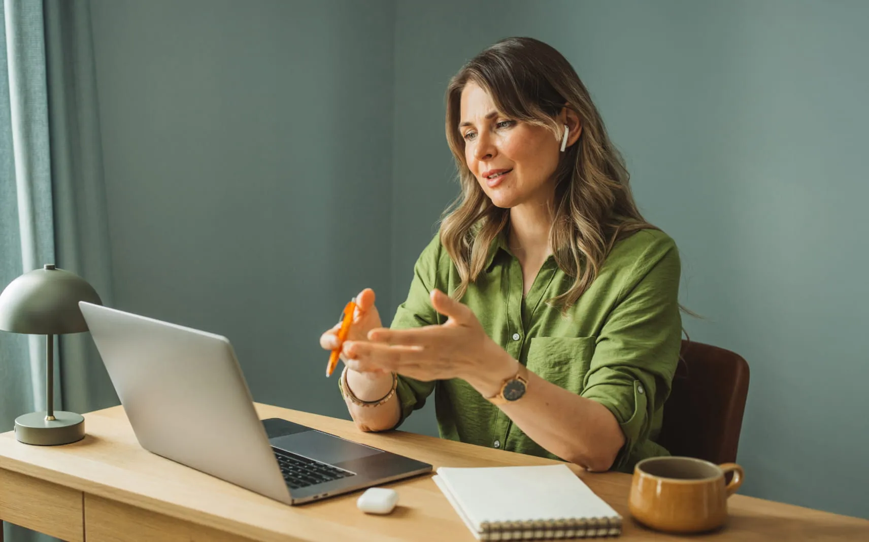 Woman wearing wireless earbuds talking on a video call at a desk with a laptop and notebook