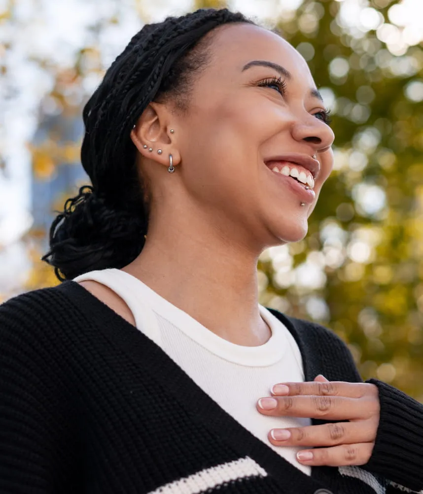 Smiling woman outdoors with braided hair, multiple ear piercings, and a hand resting on her chest, looking upward in warm sunlight.
