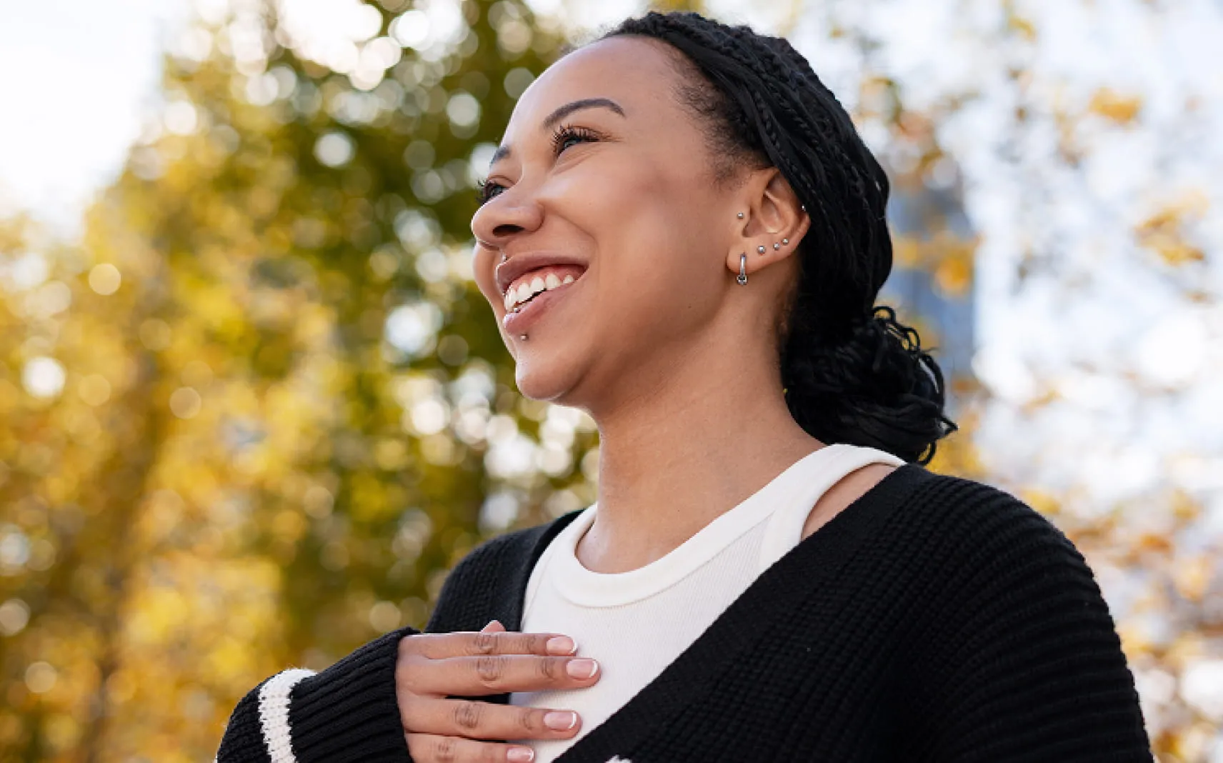 Smiling woman outdoors with braided hair, multiple ear piercings, and a hand resting on her chest, looking upward in warm sunlight.