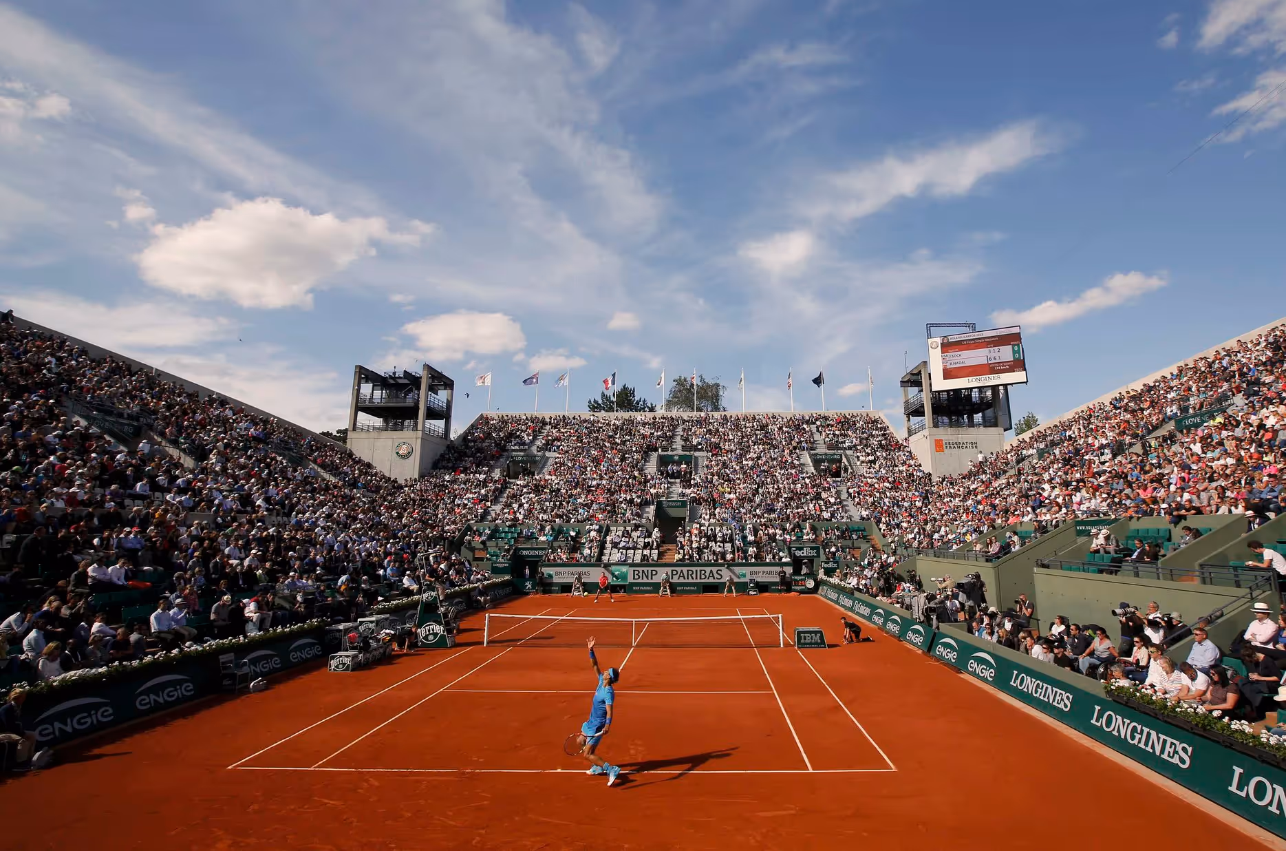 red-clay tennis court with a lot of people on the stands and a ennis player dressed in blue serving