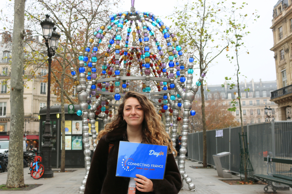 Guide holding a blue sign saying "Dayin" and "Connecting France" in front of a big and colorful glass-bubble modern sculpture.