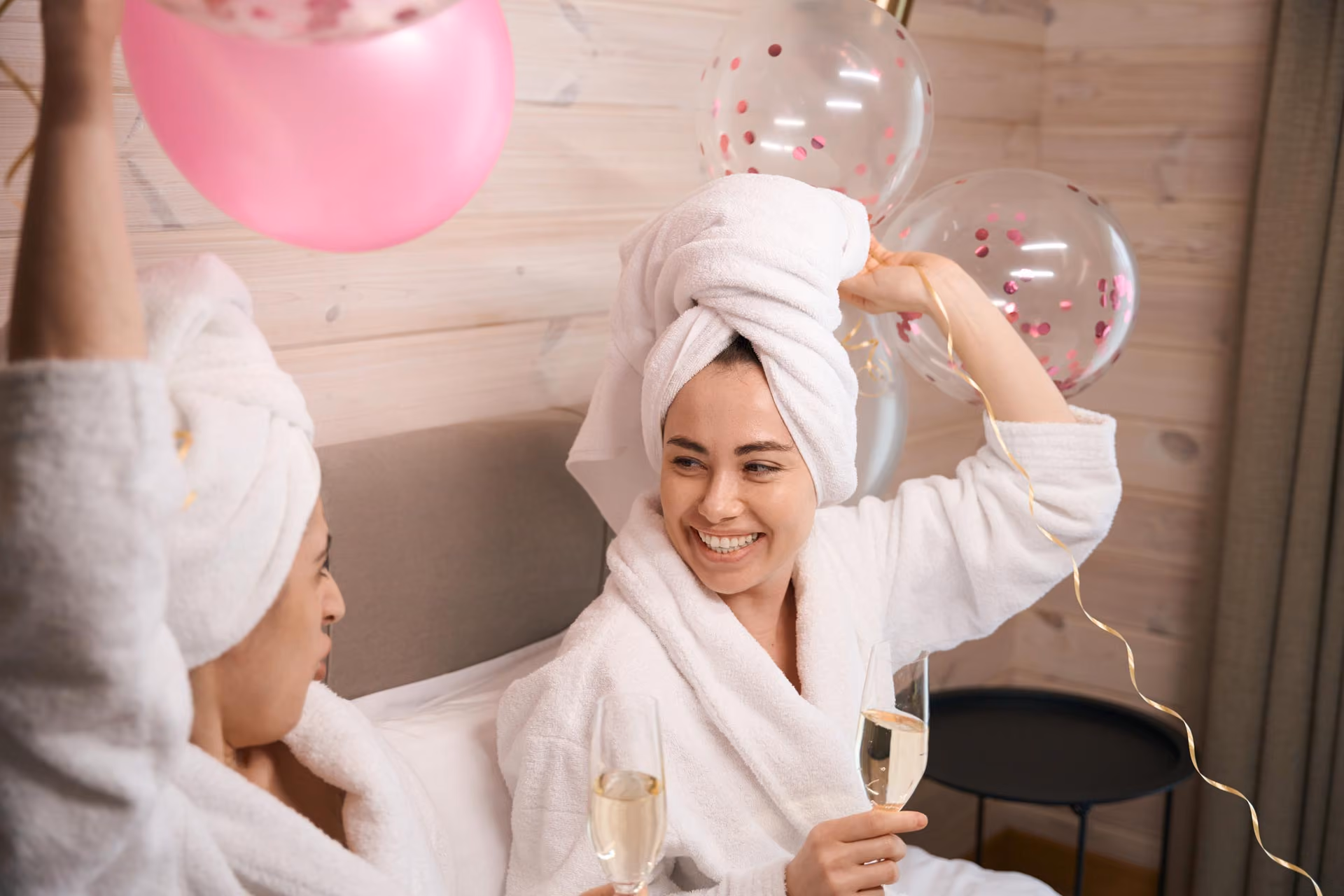 Two women in white bathrobes and towels holding champagne glasses and balloons, smiling in a cozy spa room.