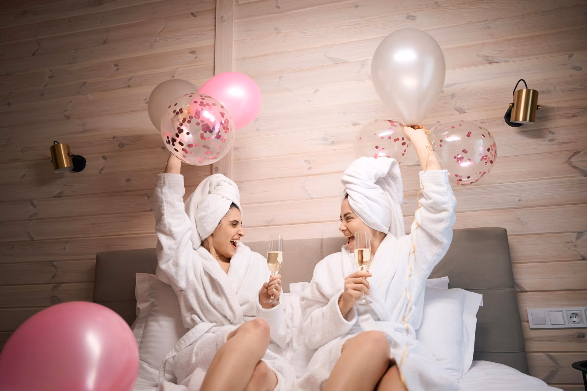 Two women in white robes with hair towels hold balloons and champagne glasses on a spa event location