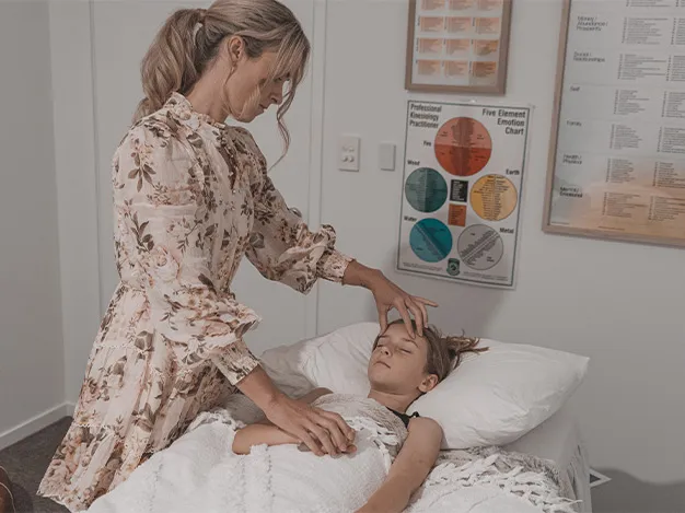 A woman in a floral dress tending to a child lying on a medical examination table. The room appears to be a medical office with informational posters on the wall.