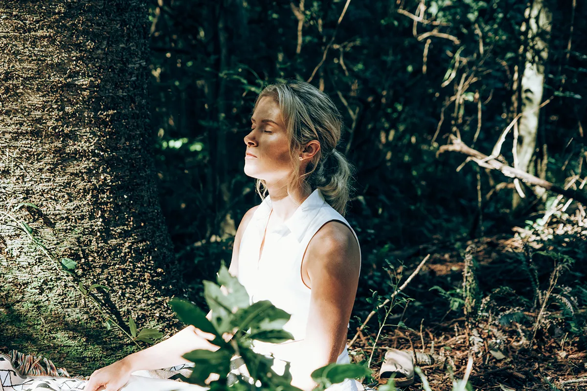 A blonde woman is seated in a meditative pose, leaning against the textured bark of a large tree.