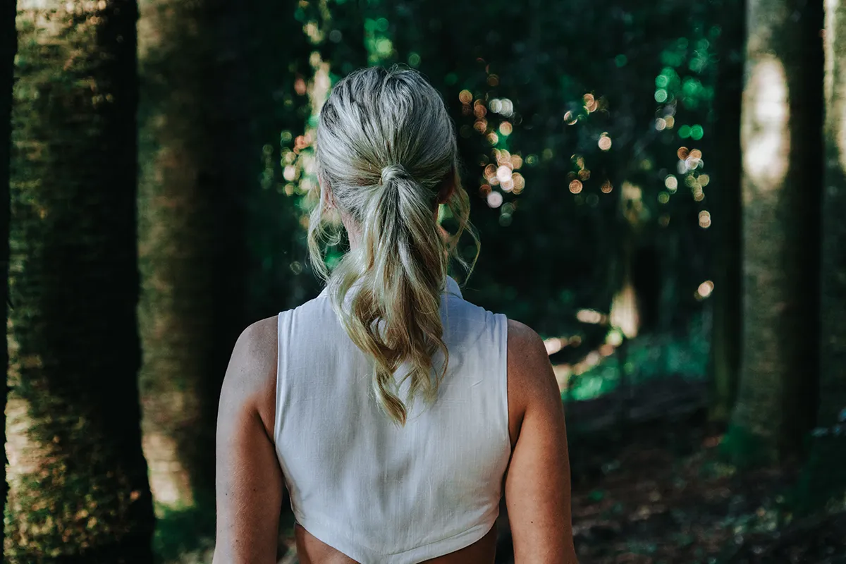 A woman with blonde hair in a low ponytail, wearing a white sleeveless top, is seen from behind in a dark, sun-dappled forest.