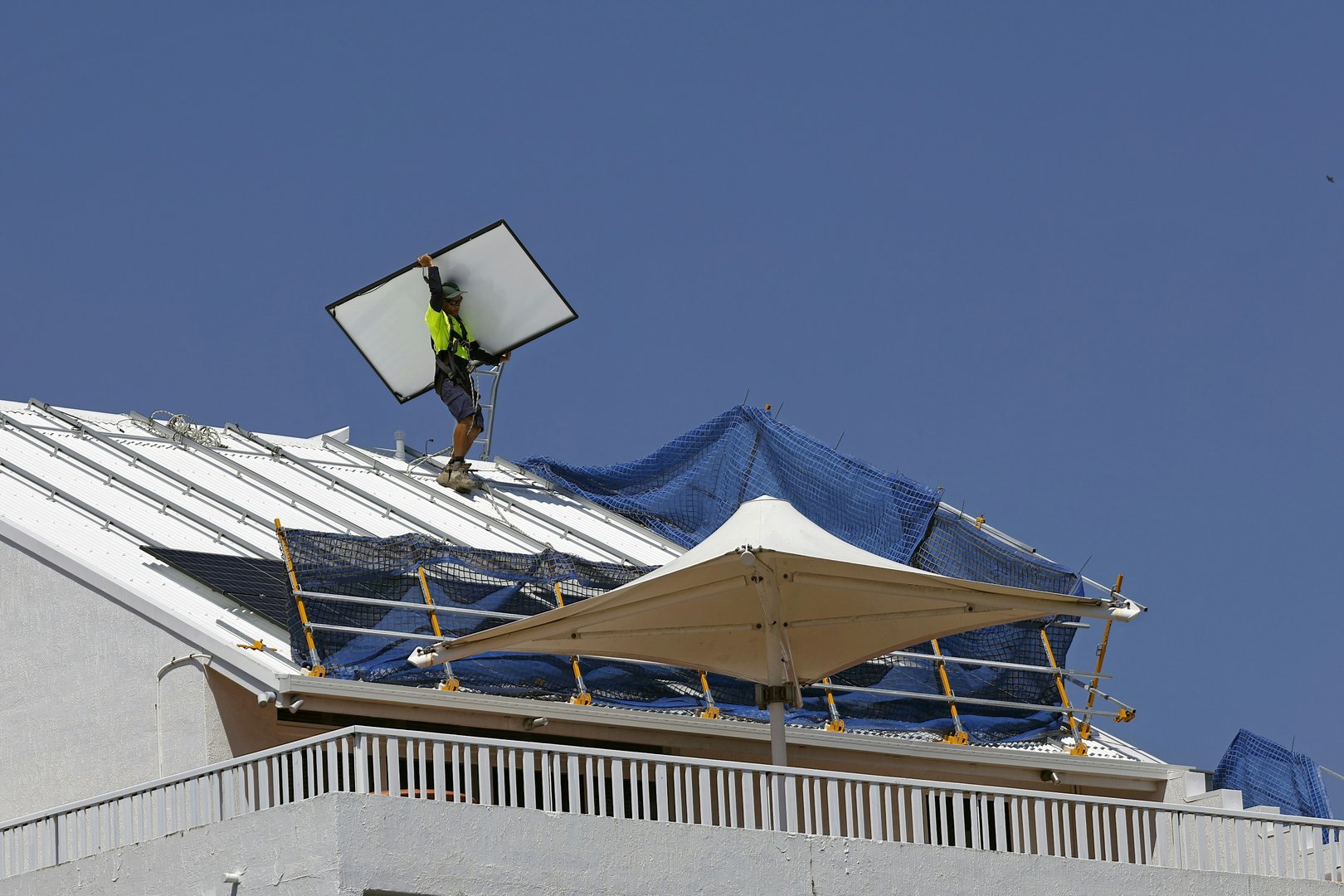 An installer in a hi-vis shirt carries a solar panel up a metal roof fitted with edge protection.
