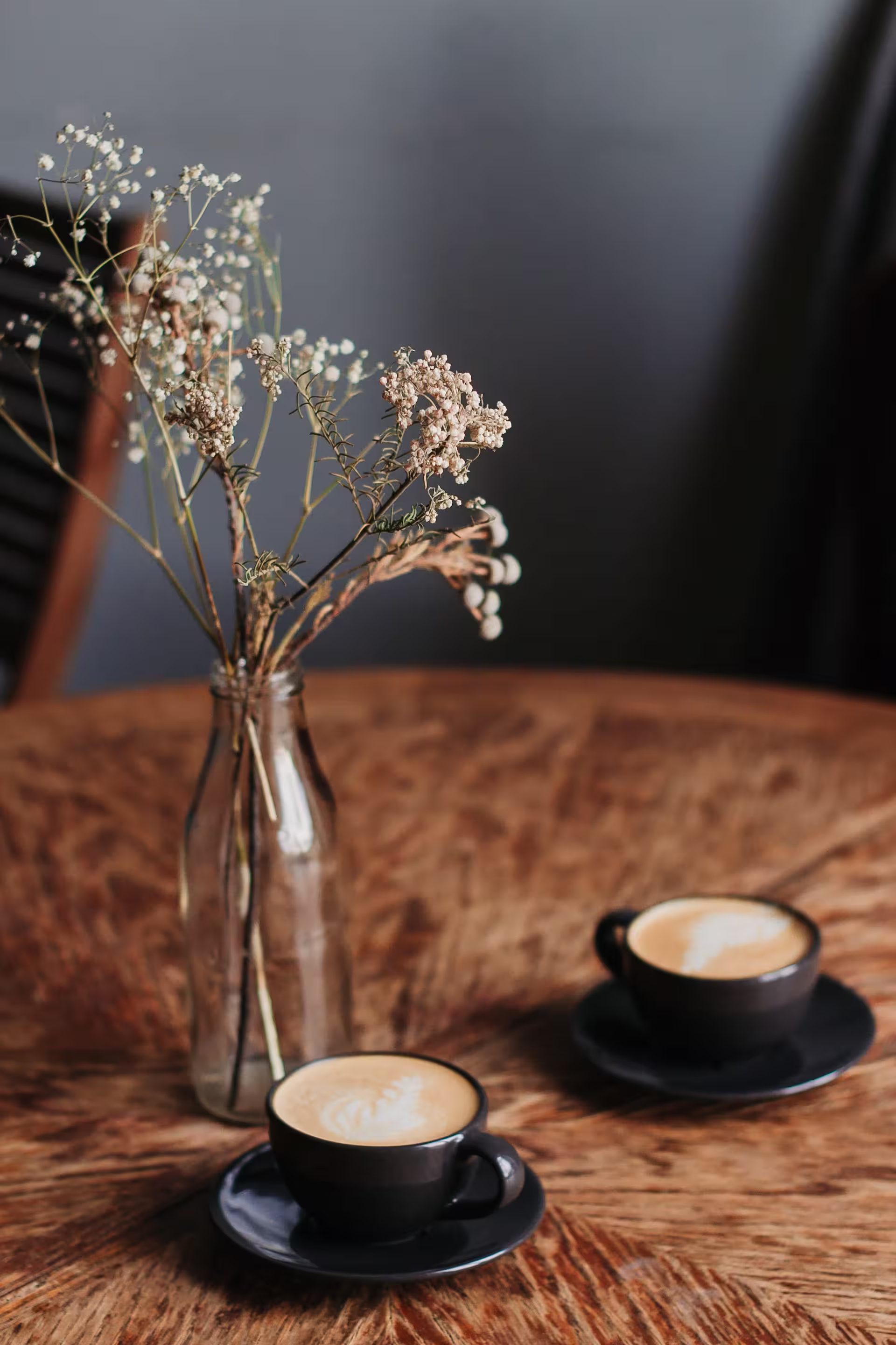 Two black coffee cups with latte art on a wooden table next to a glass bottle holding dried flowers.
