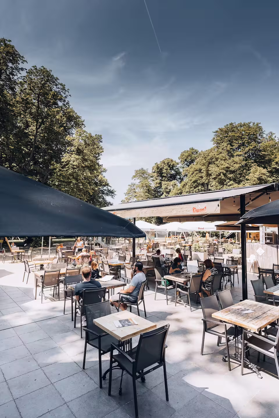 Outdoor café seating area with wooden tables and black chairs under large umbrellas, surrounded by green trees and a clear sky.