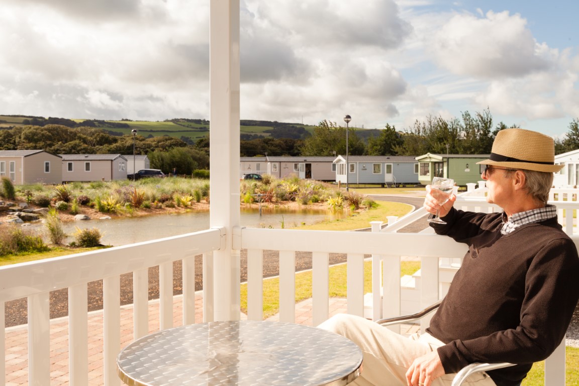 Elderly man sitting on the veranda of a luxury static caravan drinking a glass of white wine and taking in the scenic views