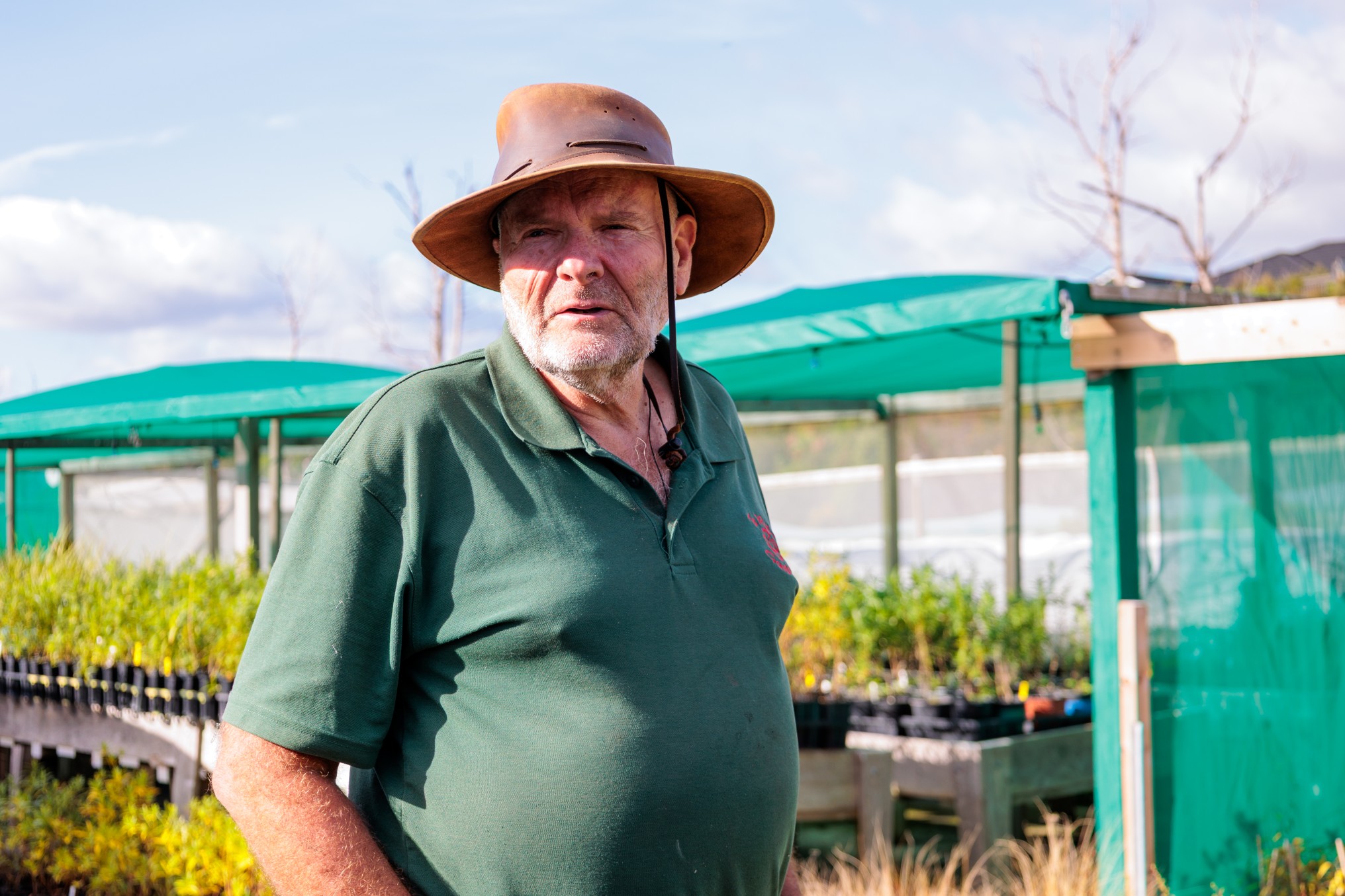 Steve Wilson wearing a wide-brimmed hat standing among rows of native seedlings under green shade cloth in an outdoor nursery.