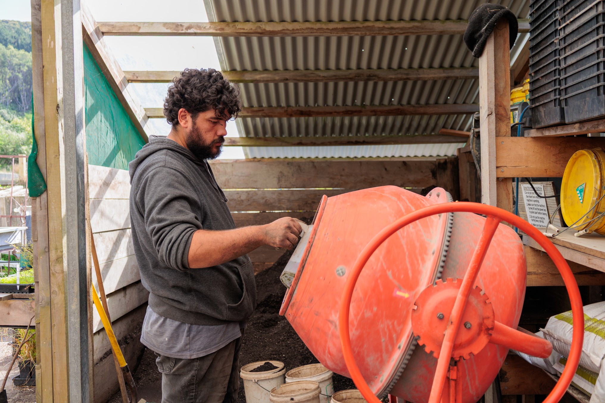 Person standing in a nursery shed pouring soil into a cement mixer, with buckets, tools, and potting supplies arranged nearby.