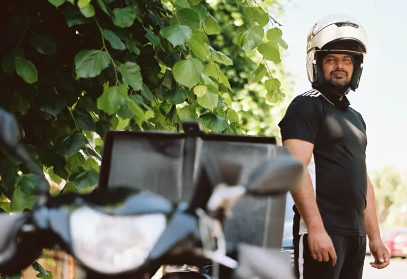Man wearing a white helmet and black sportswear standing next to a motorbike with green leafy background.