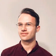 Young man with short brown hair and glasses wearing a dark red shirt, looking slightly to the side against a plain light background.
