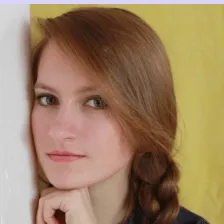 Portrait of a young woman with long brown hair styled in a loose braid, resting her chin on her hand against a yellow background.