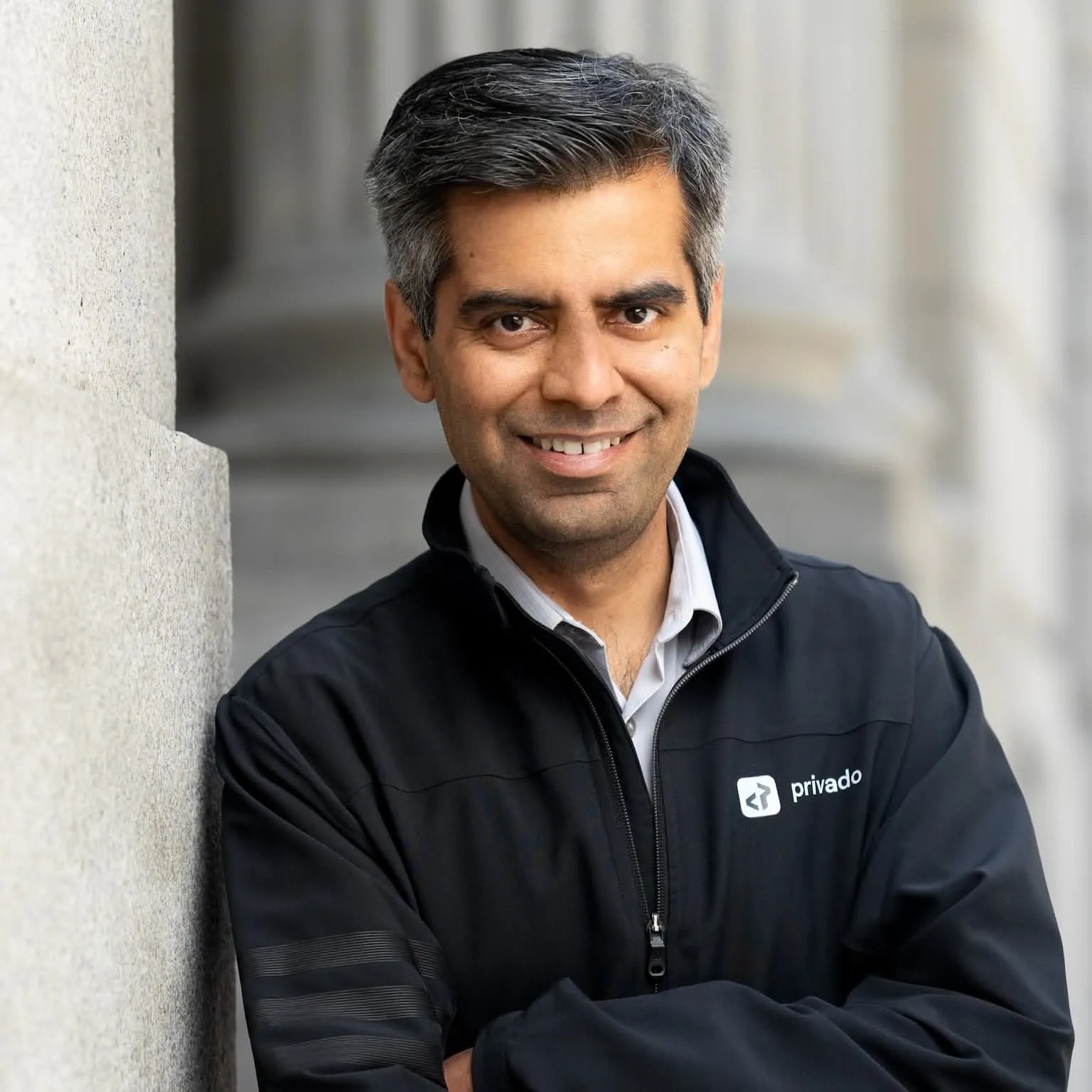 Man with salt-and-pepper hair wearing a black privado jacket, smiling with arms crossed against a stone wall.