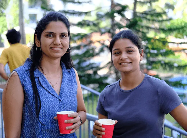 Two women smiling outdoors holding red cups with a background of green trees and a blurred person in yellow.