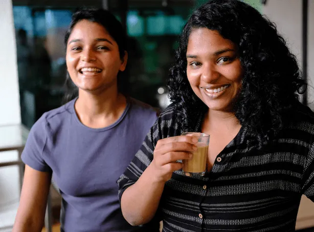 Two women smiling indoors, one holding a glass of tea or coffee.