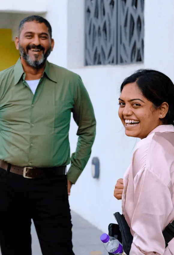 Smiling man in green shirt and smiling woman in light pink blouse with water bottle outside near white wall with window.
