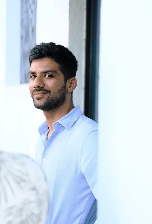 Smiling man with short dark hair and beard wearing a light blue button-up shirt, leaning against a wall.