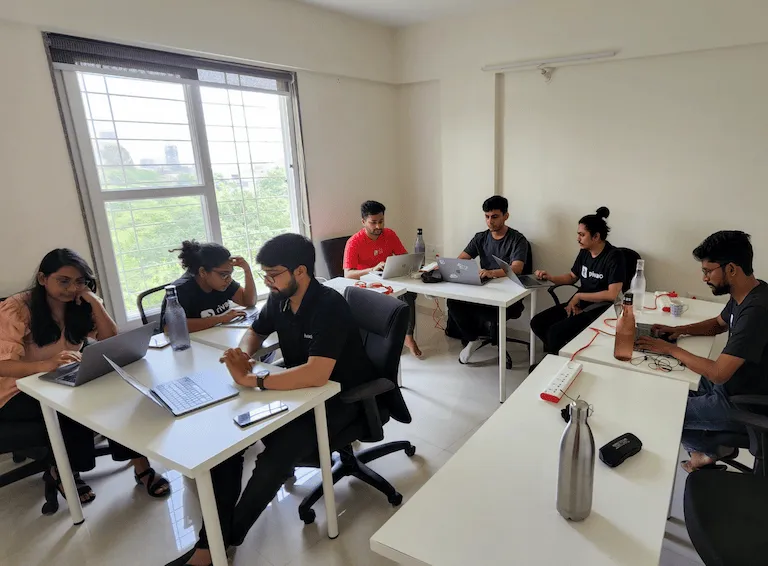 Seven people working on laptops in a bright office room with white desks and a large window showing greenery outside.