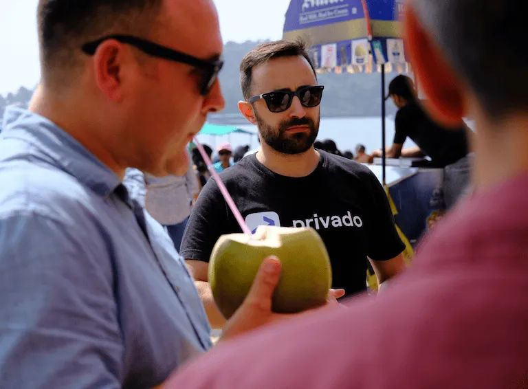 Man wearing sunglasses and a black Privado t-shirt at an outdoor event, with another man drinking from a coconut using a straw in the foreground.