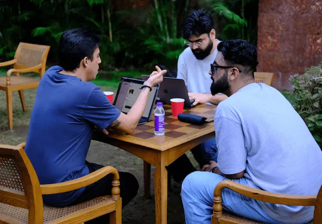Three men sitting around a wooden table outdoors, two using laptops and one gesturing while talking.