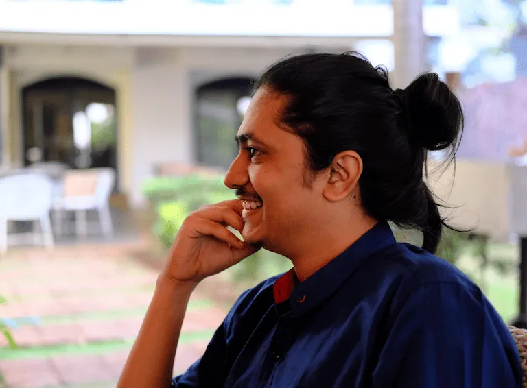 Side profile of a smiling man with a man bun, resting his chin on his hand while sitting outdoors.