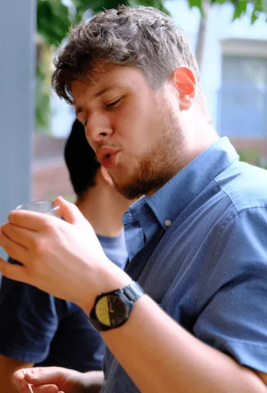 Young man with short curly hair and beard wearing a blue shirt and watch, holding and looking at a small cup.