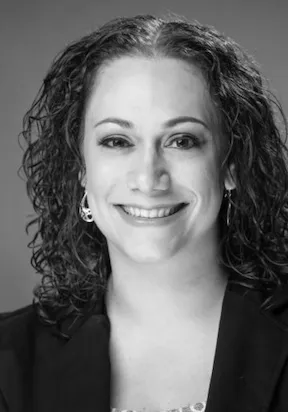 Black and white portrait of a smiling woman with curly hair wearing earrings and a dark blazer.