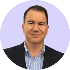 Man in a dark blazer and blue striped shirt with a neutral expression against a light purple background.