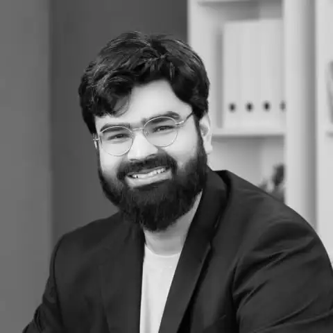 Smiling bearded man with glasses wearing a dark blazer and white shirt in an office setting.