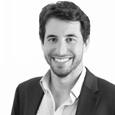 Smiling man with short dark hair and beard wearing a suit jacket and open-collar shirt on a white background.